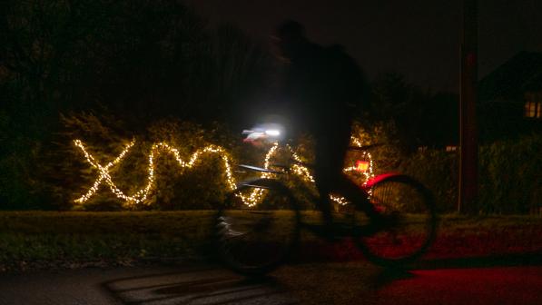 XMAS is spelled out in Christmas lights in a hedge, with a person in silhouette on a bike cycling past standing on the pedals