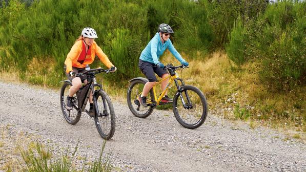 Two people are riding along a gravel track in the Scottish Highlands. They are on mountain bikes and are wearing MTB kit and cycling helmets