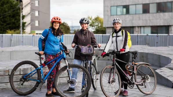 Three women stand together with bikes on an urban plaza, smiling at the camera