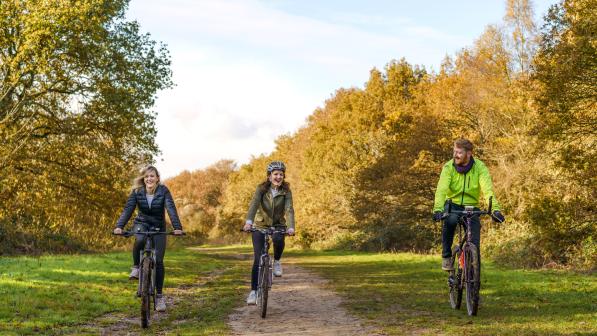 Three people are cycling along a dirt track. It's autumn and the trees are all yellow. The people are wearing long-sleeved jackets and leggings. They are smiling
