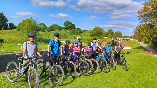 A line of people all holding mountain bikes and wearing cycling kit and helmets are standing on grass next to a canal. It looks sunny and the sky is blue