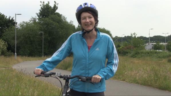 A smiling woman wearing a blue cycling helmet and a blue Adidas jacket stands with her hands on the handlebars of a bicycle. She is on a paved path surrounded by grass and trees, with streetlights and a few buildings in the background.