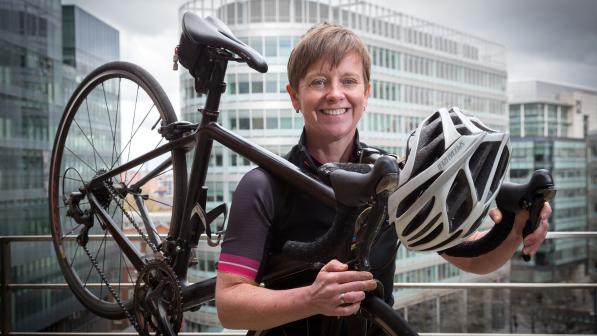 A woman standing on what looks like a balcony in an office building in a city with lots of office blocks behind her. She is wearing cycling kite and holding a black road bike and white Specialized cycling helmet.