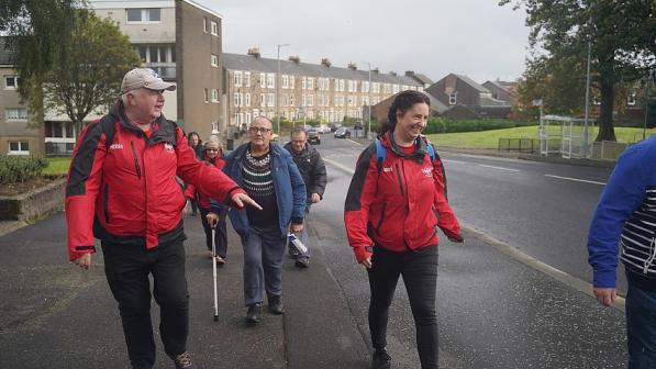 A group of people participating in a community walk along a residential street. Two leaders in red jackets are smiling and engaging with the group, which includes older participants, some using walking aids. The background features rows of terraced houses and a green open space, creating a welcoming and inclusive atmosphere of activity and connection.