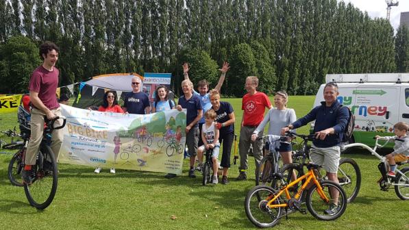 A group of people smiling and posing with bikes at an outdoor cycling event on a sunny day. Some are holding a colourful “Big Bike Revival” banner featuring cartoon bikes and logos, while others stand beside or ride bikes, including children and adults. A man at the front left is performing a wheelie on a bike. A branded van and tent with "Avanti Cycling" signage are visible in the background, along with tall trees lining the park.