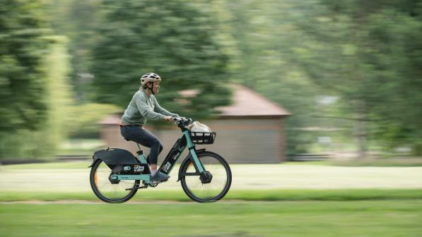 A woman is riding an e-bike in a park. It's green and black and has a Making cycling e-asier logo on it. There's a basket on the front with some shopping in it. The rider is wearing normal clothes and a cycle helmet. She is smiling