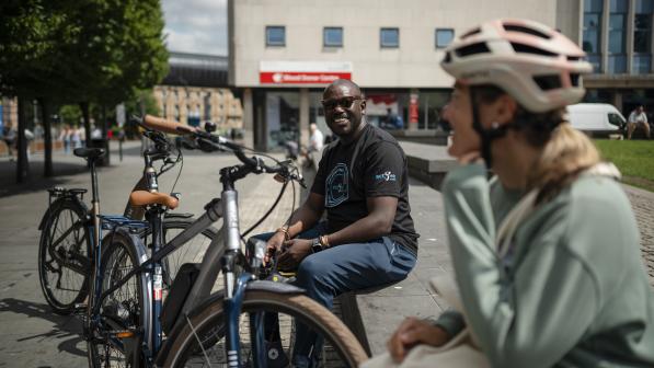  A man in jeans and T-shirt and a woman in a sweater and cycling helmet are sitting on a wall in an urban setting. Two bikes are leaning against the wall with them