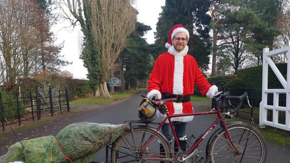 A cyclist dressed as Santa standing next to a bike with a Christmas tree in tow. 
