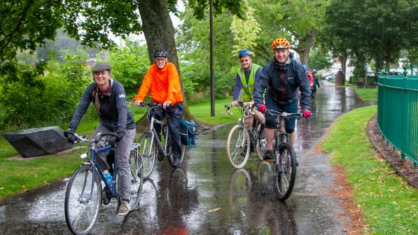 A group of cyclists riding together on a rainy day through a park, wearing weather-appropriate gear such as helmets, rain jackets, and reflective vests. The path is lined with green trees and wet pavement reflecting the vibrant colours of their clothing, creating a lively and determined atmosphere.
