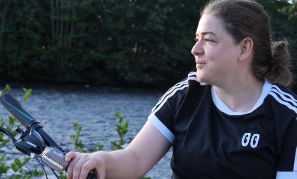 A woman in a navy blue t-shirt with white stripes sits on a bicycle by a lake while holding on to her handlebars 