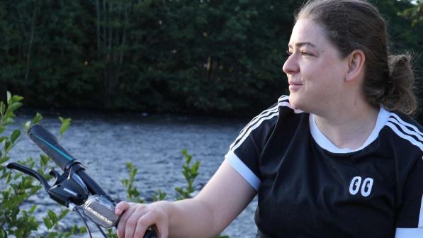 A woman in a navy blue t-shirt with white stripes sits on a bicycle by a lake while holding on to her handlebars 