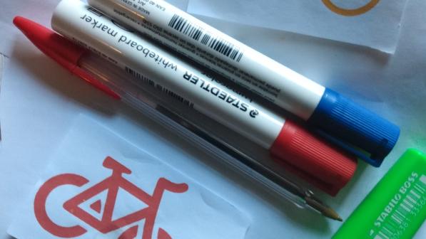 Two board markers, a red pen, a green pen and red and yellow line pictures of bicycles on a white backdrop