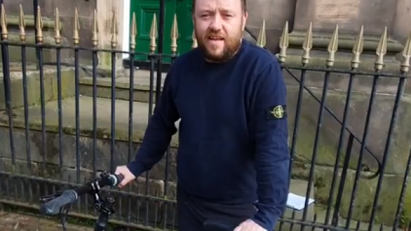 Freezeframe from a video of a man in a navy pullover standing behind a bicycle 