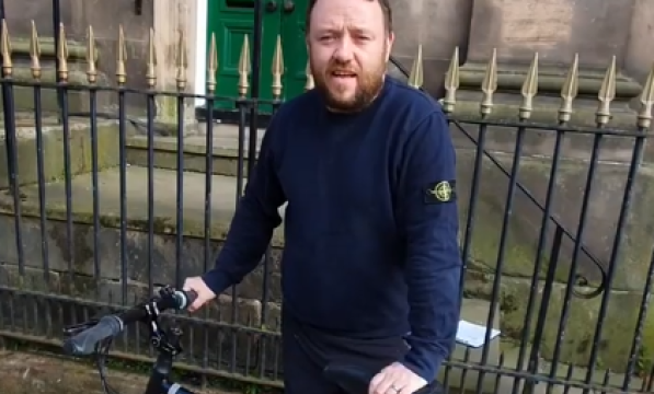 Freezeframe from a video of a man in a navy pullover standing behind a bicycle 