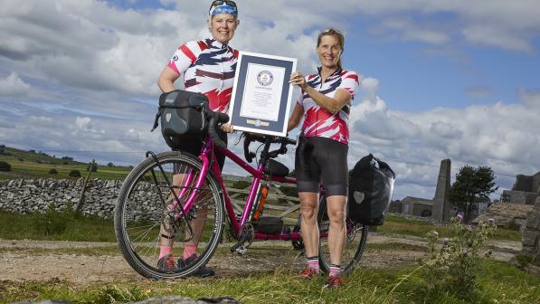 Two women in cycling kit stand either side of a pink tandem bicycle holding a framed certificate 