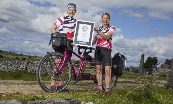 Two women in cycling kit stand either side of a pink tandem bicycle holding a framed certificate 