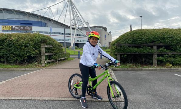 A young boy poses on a bicycle outside a large football stadium 