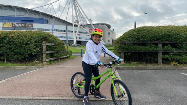 A young boy poses on a bicycle outside a large football stadium 