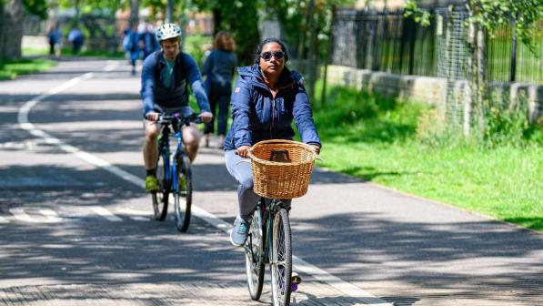 Two people are cycling on a dedicated cycle path alongside a pedestrian path. One is on a mountain bike, the other on a hybrid with a wicker basket on the front. They are both wearing normal clothes.