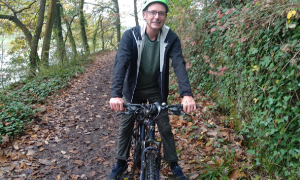 A middle-aged man wearing a green helmet sits astride a bicycle in a woodland setting 