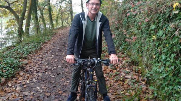 A middle-aged man wearing a green helmet sits astride a bicycle in a woodland setting 