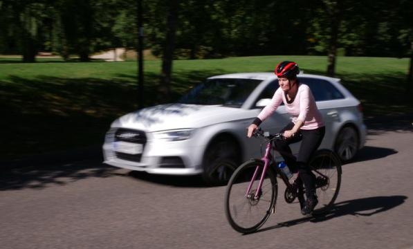 A car accelerates past a female cyclist wearing a helmet