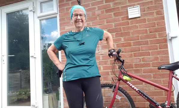 A woman wearing a azure fabric cycling top stands next to her bicycle on her patio 