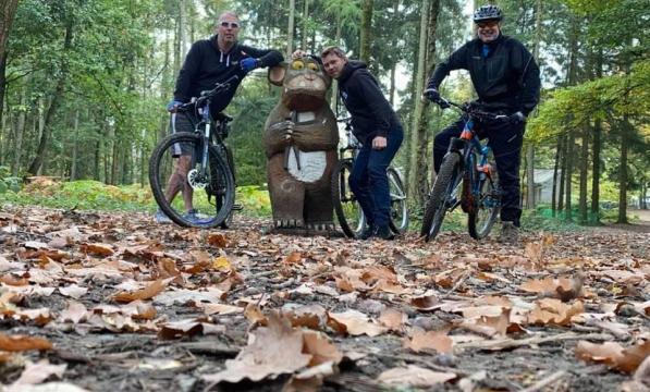 Three men on mountain bikes in a forest trail stand by a Gruffalo statue 