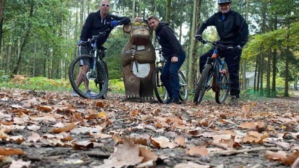 Three men on mountain bikes in a forest trail stand by a Gruffalo statue 