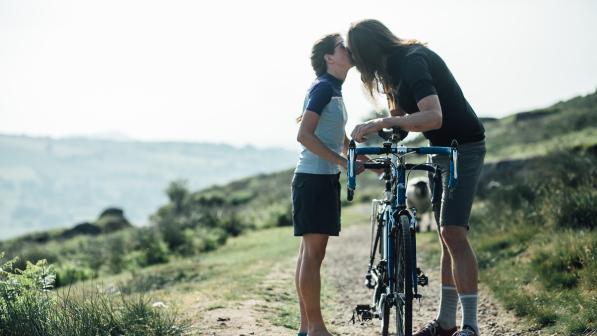 Two people are standing on an off-road track holding a bike in between them, leaning over it and kissing