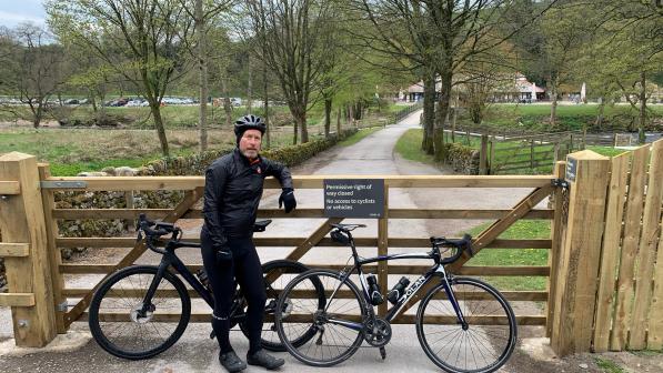 A cyclist dressed in black with two black bicycles stands in front of a barred wooden gate 