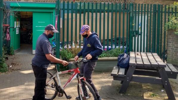 Two men stand either side of a red and white bicycle outside a green door and a set of high railings. They are standing to the left of the frame, a picnic bench is on the right. 