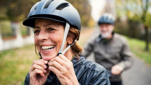 A woman adjust the chin straps of her helmet. A man can be seen out of focus in the back of the shot.