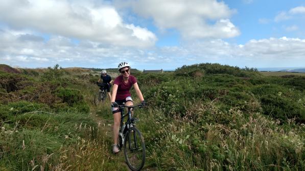 A young woman cycles towards us among a moorland vista followed by a young man