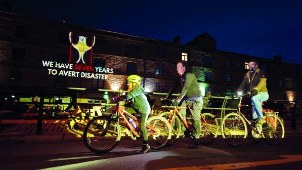 Two men and a young girl cycle on bikes in front of a lit up building