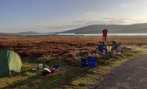A bicycle is propped up against a signpost next to a pitched tent in front of a loch and mountains in the background