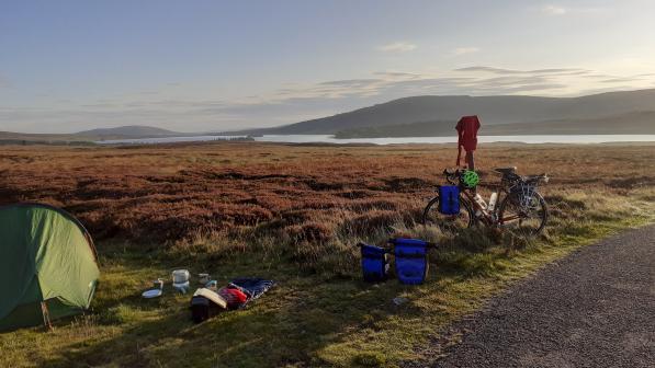 A bicycle is propped up against a signpost next to a pitched tent in front of a loch and mountains in the background