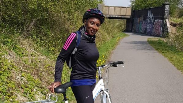 A woman is standing astride a hybrid biked on a paved path. She is wearing cycling kit and helmet