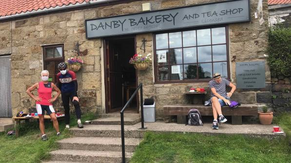 A man and woman wearing face masks pose in their cycling gear outside of a bakery. To the left of them is a lady sat on a bench tucking into her lunch