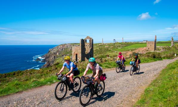 A group of cyclists ride towards camera on a gravel track along the Cornish coast. The background is scattered with tin mines