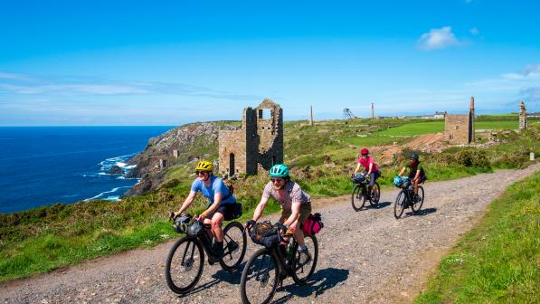A group of cyclists ride towards camera on a gravel track along the Cornish coast. The background is scattered with tin mines