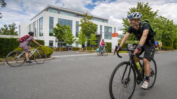 A man cycles towards camera on a road bike, he is wearing shorts and a t-shirt and carrying a backpack. In the background two women are cycling past a large office building