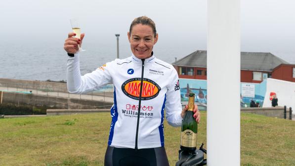 A woman holds up a glass of champagne as she stands underneath the John O' Groats sign in Scotland. She's wearing cycling jersey and bibs