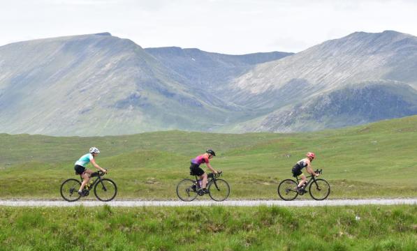 Three women road cycling in single file with a hazy mountain landscape in the background