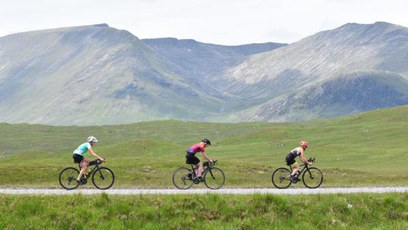 Three women road cycling in single file with a hazy mountain landscape in the background
