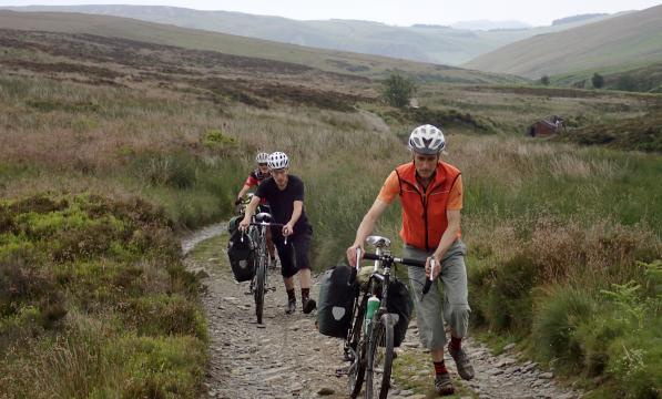 Three men on foot pushing their bikes up hill. Behind them is a beautiful lush green valley, a small abandoned building in the background