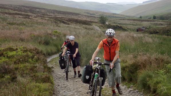 Three men on foot pushing their bikes up hill. Behind them is a beautiful lush green valley, a small abandoned building in the background