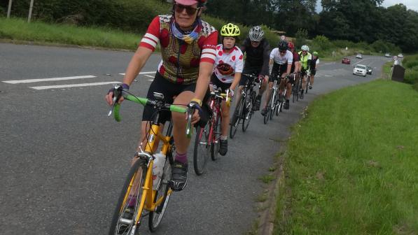 A line of seven cyclists of mixed ages and sexes cycle in a line along a busy road