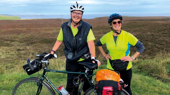 Two women stand behind a Dawes Galaxy touring bike that's loaded with panniers. They are both wearing helmets and high vis tops and smiling broadly at the camera. In the background is a coastline covered in heather.