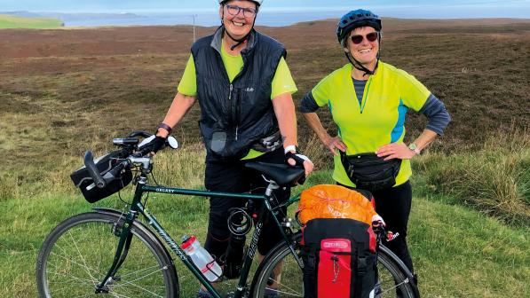 Two women stand behind a Dawes Galaxy touring bike that's loaded with panniers. They are both wearing helmets and high vis tops and smiling broadly at the camera. In the background is a coastline covered in heather.